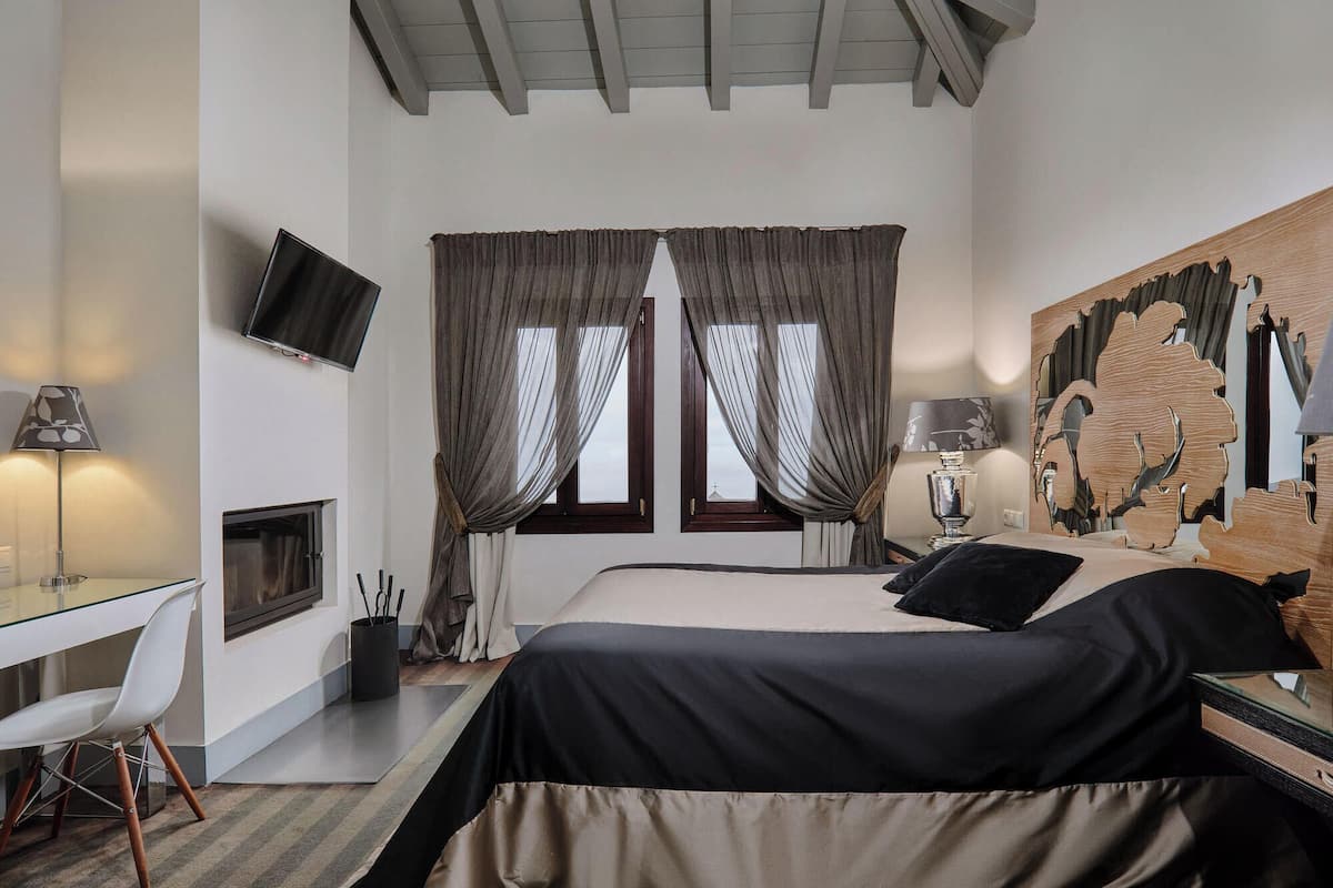 A modern bedroom with a bed, two windows with curtains, a flat-screen TV, a desk with a lamp, and a decorative wall piece.