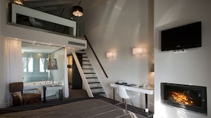 A modern bedroom with a large bed, a desk, a chair, a fireplace, and a staircase leading to an upper level. - Despotiko Hotel (Volos)