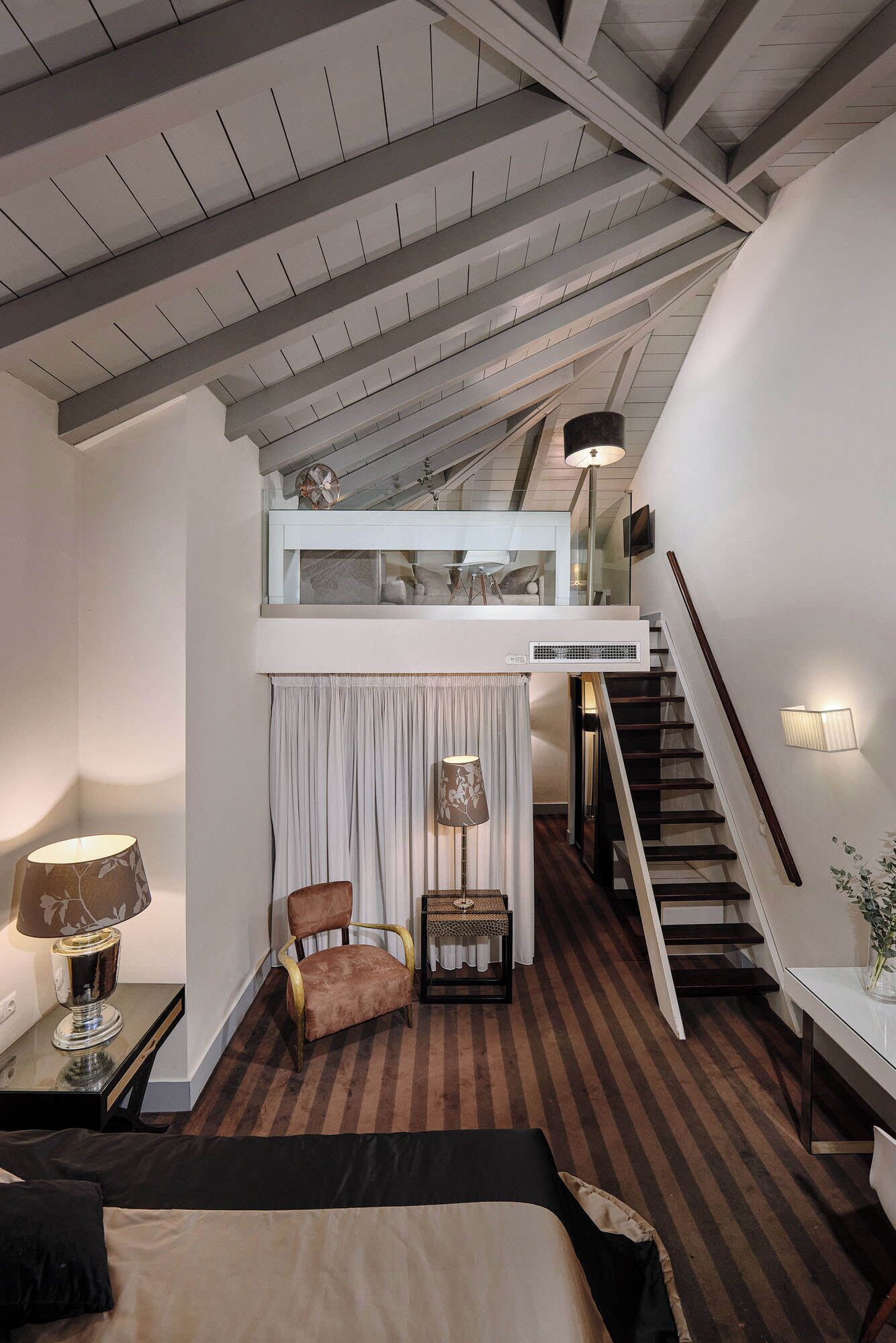 Une chambre à coucher comprenant un lit, une chaise, une petite table, une lampe, un escalier et un plafond avec des poutres apparentes.