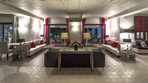 A spacious living area with a central sofa, a coffee table, and a dining table. There are two side tables with lamps, and a view into the dining area through a glass door. - Despotiko Hotel (Volos)