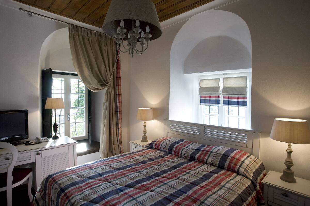 A bedroom with a bed, a desk, a chair, a television, and a window with curtains.