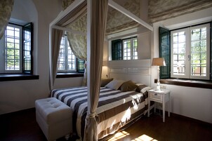 Una cama con dosel y cortinas, una mesita de noche con lámpara, y una ventana con vistas al verdor.
