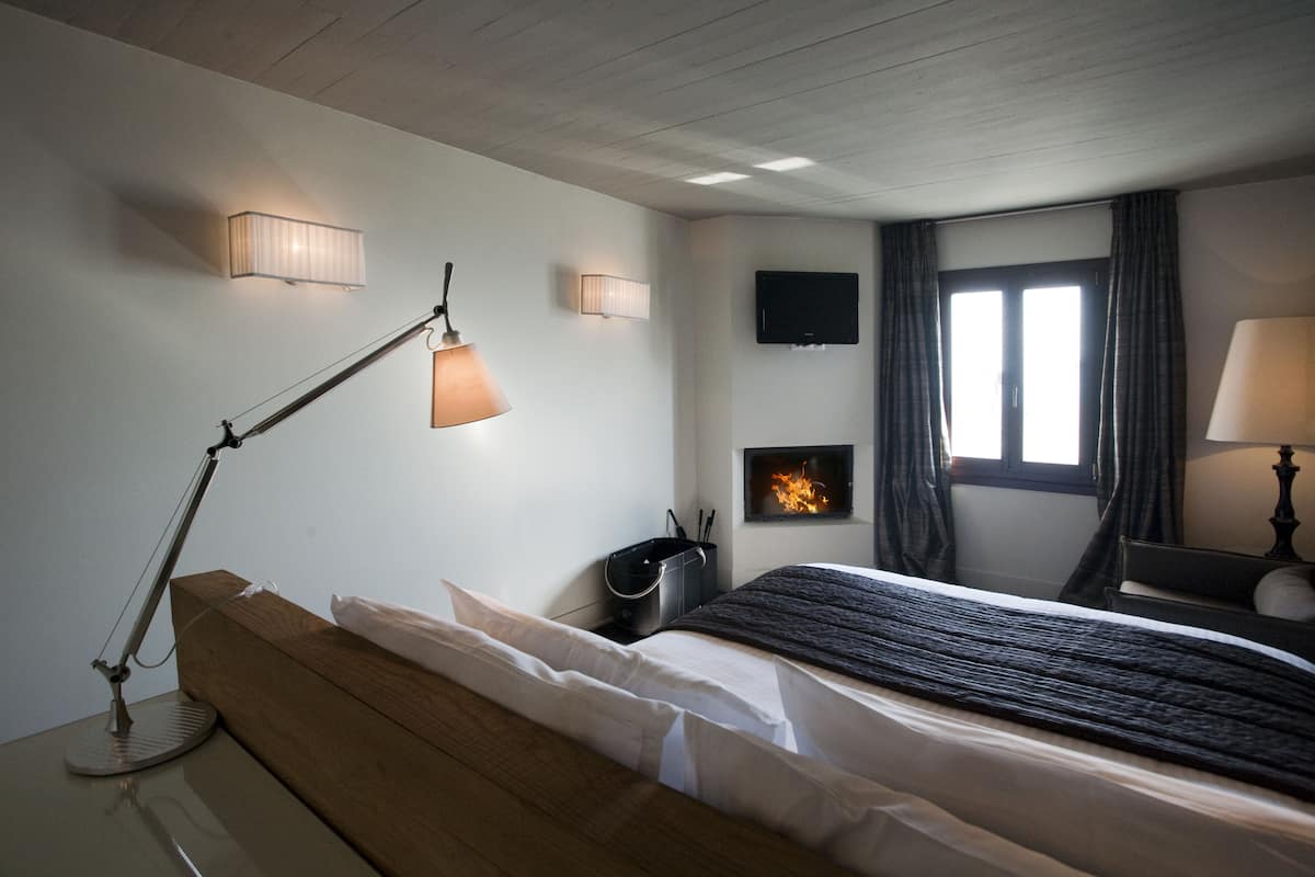 A bedroom with a wooden headboard, a bedside lamp, a flat-screen TV, a fireplace, and a window with curtains.