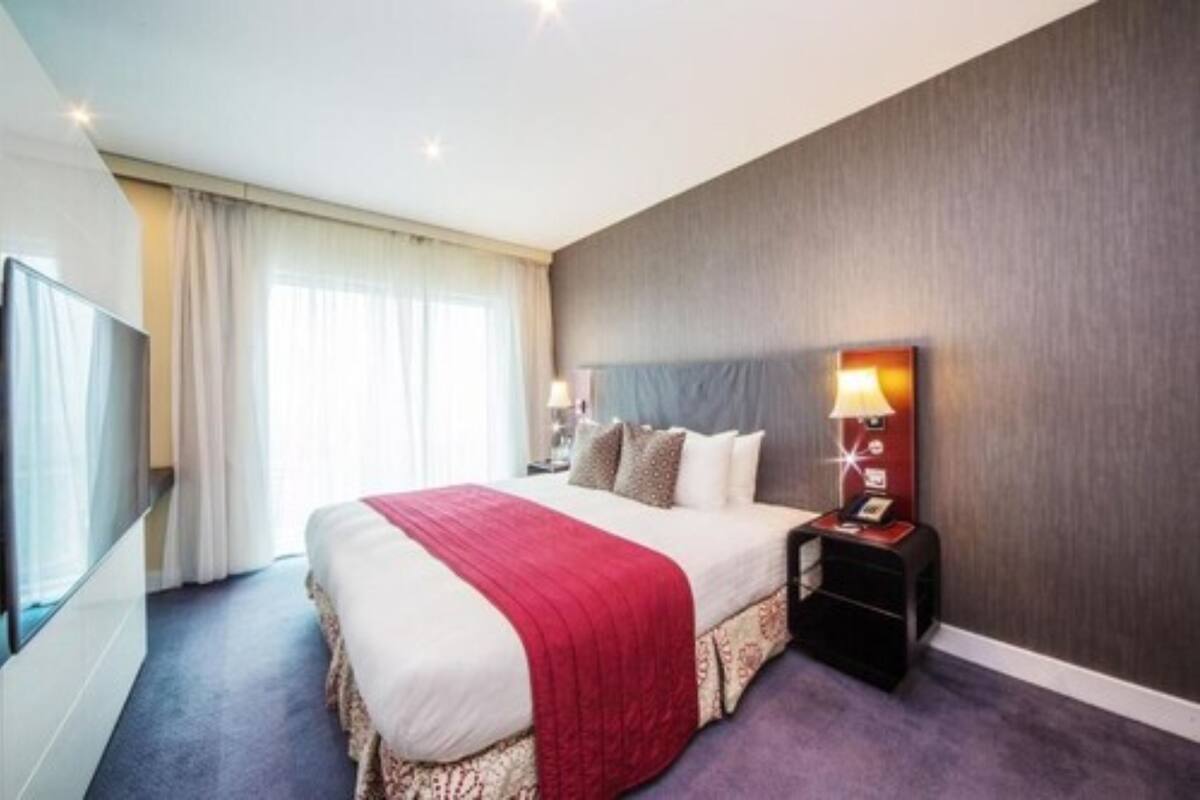 A hotel room with a large bed, a bedside table with a lamp, a TV, and a window with curtains.
