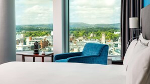 Quarto de hotel com uma janela grande, uma cama, uma poltrona azul e uma mesinha com chaleira e chávena.