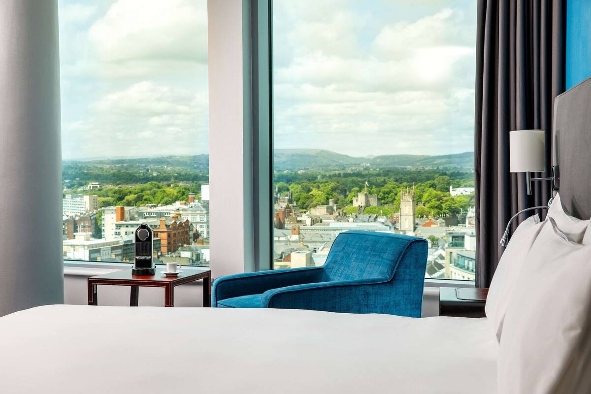 A hotel room with a large window, a bed, a blue armchair, and a small table with a kettle and cup.