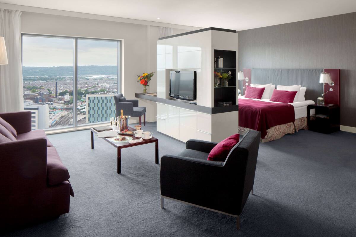A modern hotel room with a large window, a flat-screen TV, a sofa, a chair, a bed with a red blanket, and a small table with a vase and a bottle.