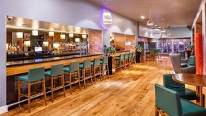 Uma área de bar com piso de madeira, banquetas e balcão com um computador e diversos frascos.