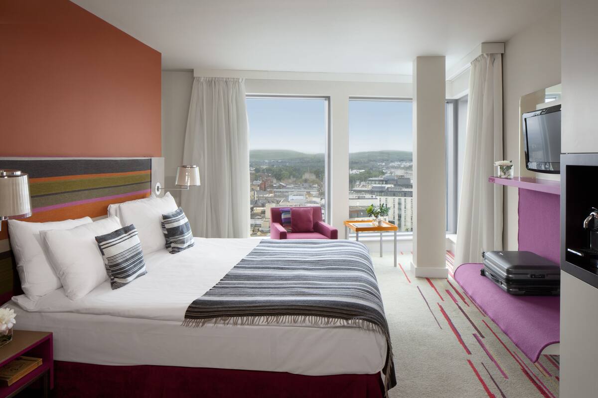 A hotel room with a large bed, a desk, a chair, a TV, and a view of the city.