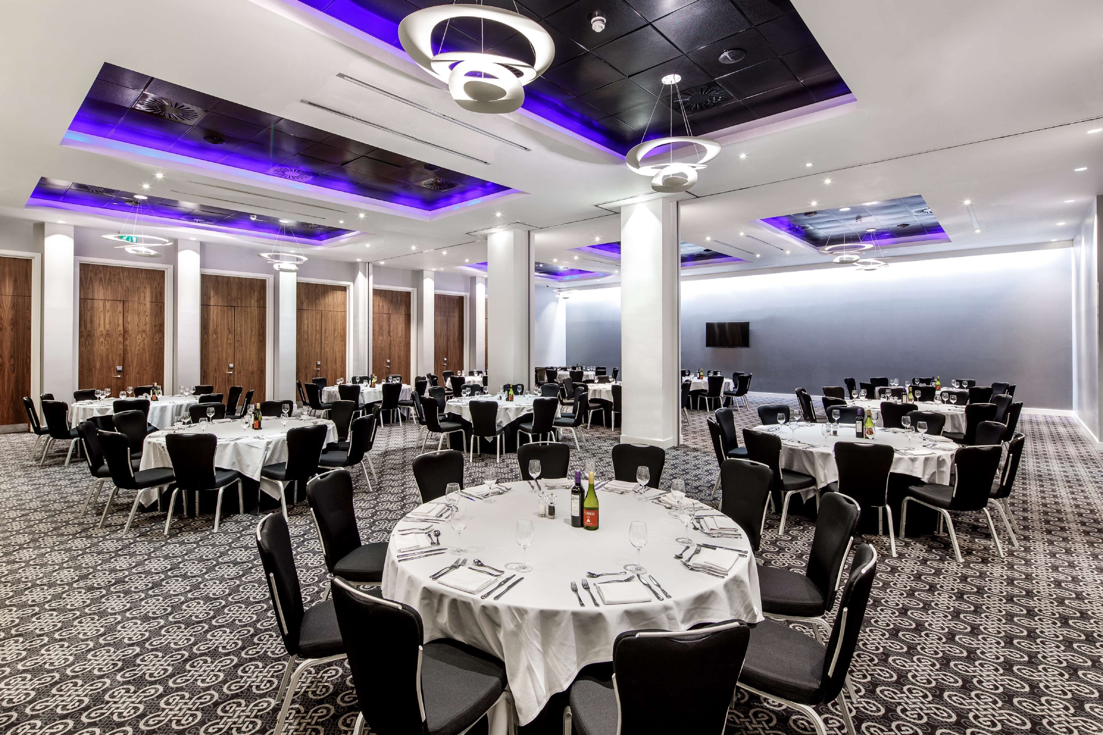 Une vaste salle de banquet avec des tables rondes dressées pour le repas, des panneaux de plafond blancs et des luminaires modernes.