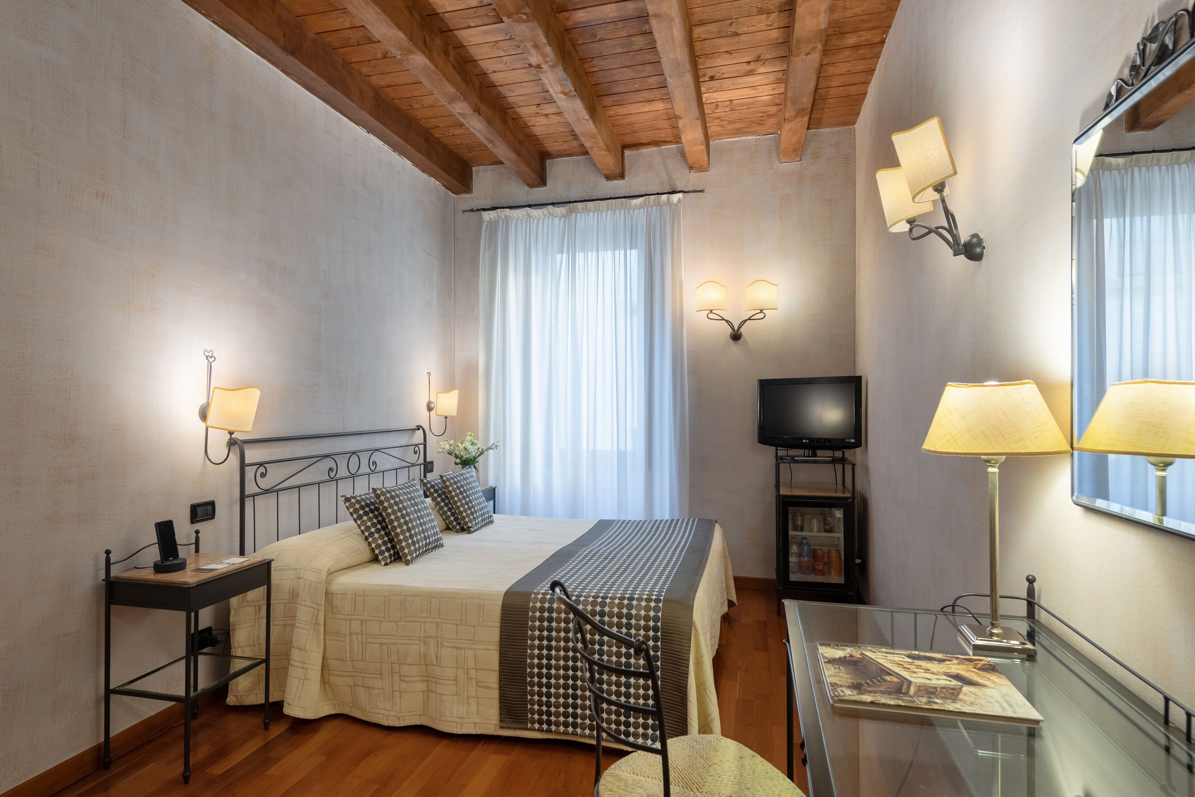 A bedroom with a bed, bedside table, lamp, TV, and a glass table with a chair.
