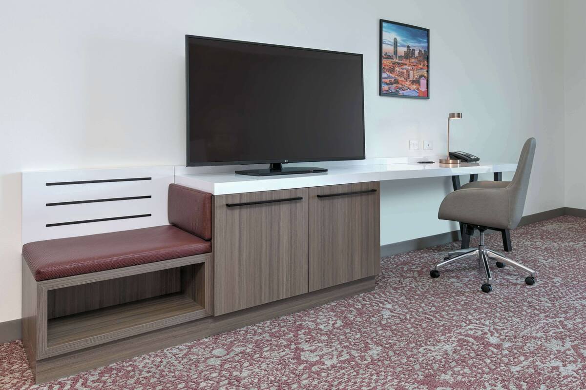 A modern office space with a large flat-screen TV, a desk, a cushioned bench, and a chair.