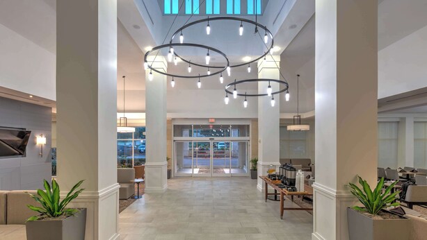 A spacious lobby with a modern chandelier, a reception area, and a view of the parking lot through glass doors. - Hilton Garden Inn Dallas/Arlington (Arlington)