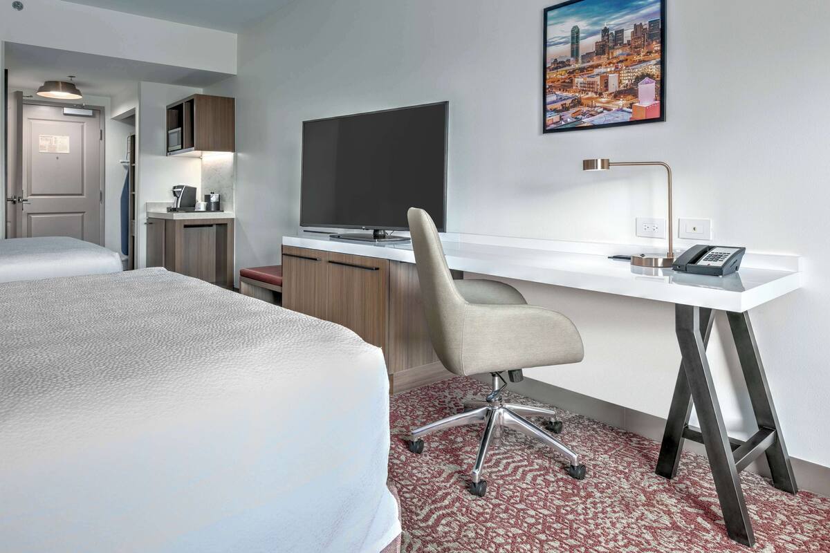 A hotel room with a bed, a desk with a chair, a television, a desk lamp, and a cityscape painting on the wall.