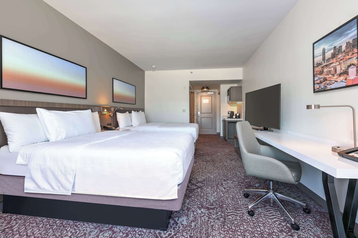 A hotel room with a large bed, a desk, and a chair. There are two paintings on the walls and a cityscape picture above the desk.