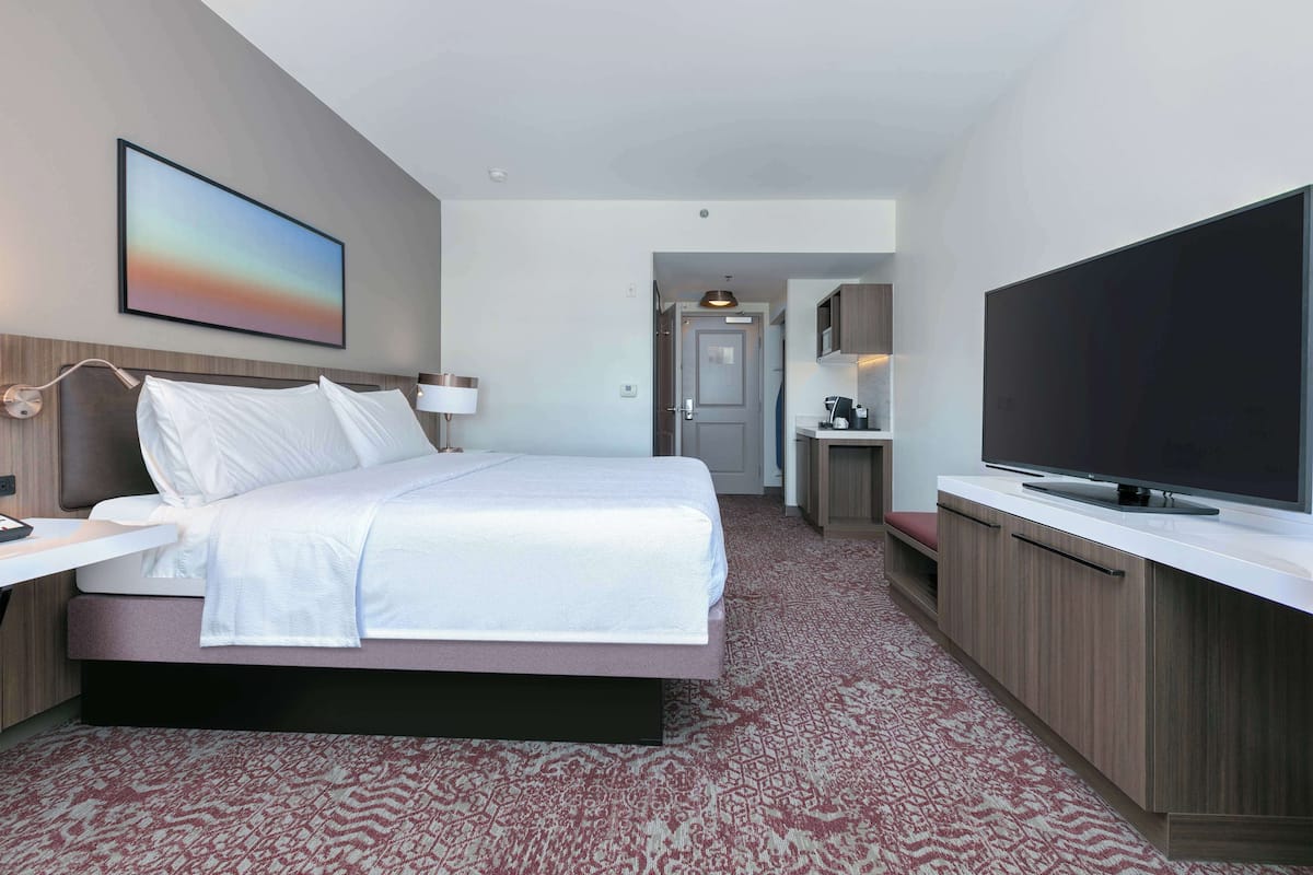 A modern hotel room with a bed, a TV, a desk, and a bathroom visible through an open door.
