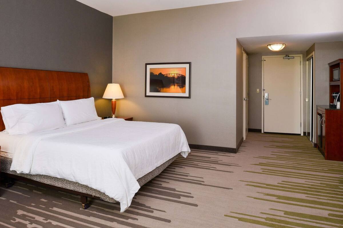 Une chambre d’hôtel avec un lit, une table de chevet, une lampe, un tableau encadré au mur et un couloir menant à une porte.