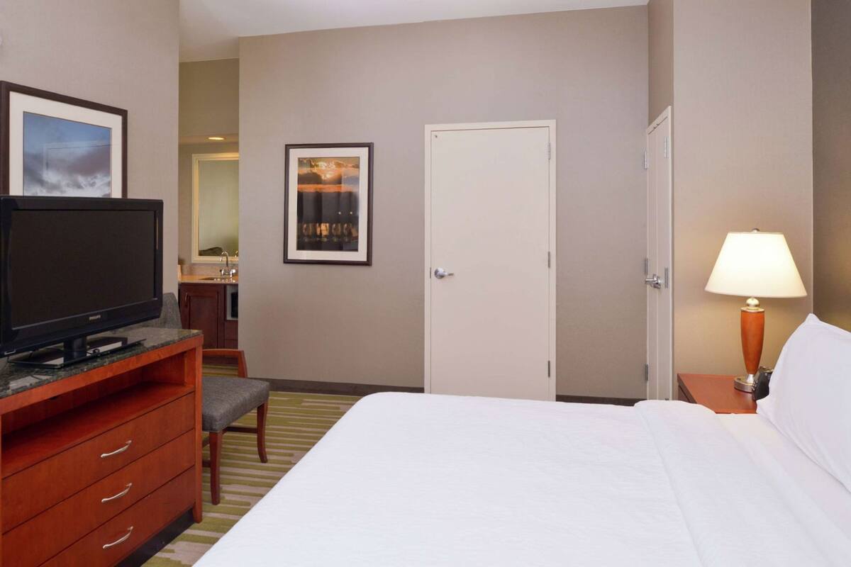 Une chambre d’hôtel avec un lit, un téléviseur posé sur un meuble en bois, un bureau avec une chaise, une porte et une lampe.