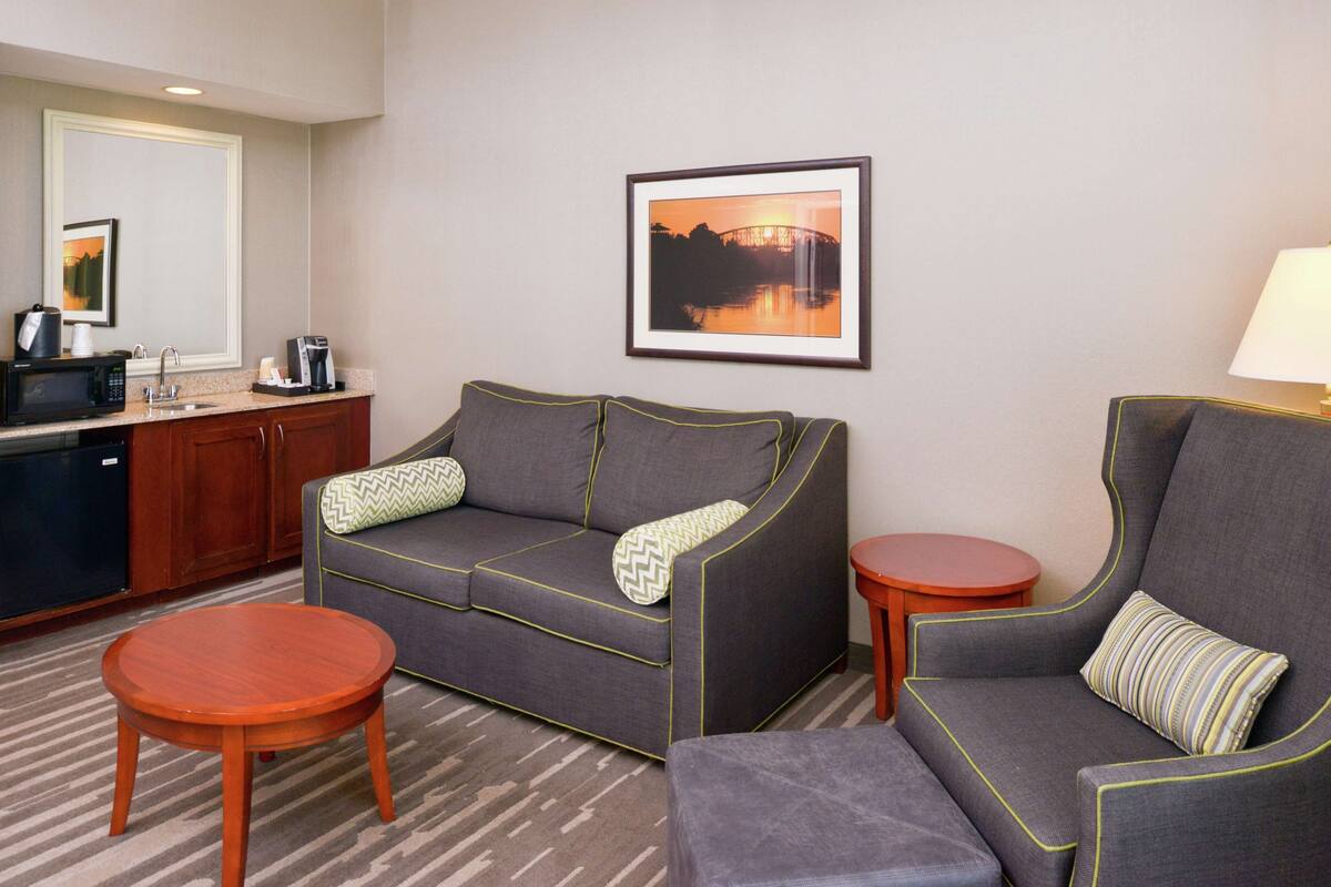 Une chambre d’hôtel comprenant un canapé, un petit divan, une table ronde en bois, une petite table d’appoint, un coin cuisine avec un four à micro-ondes et un tableau encadré au mur.
