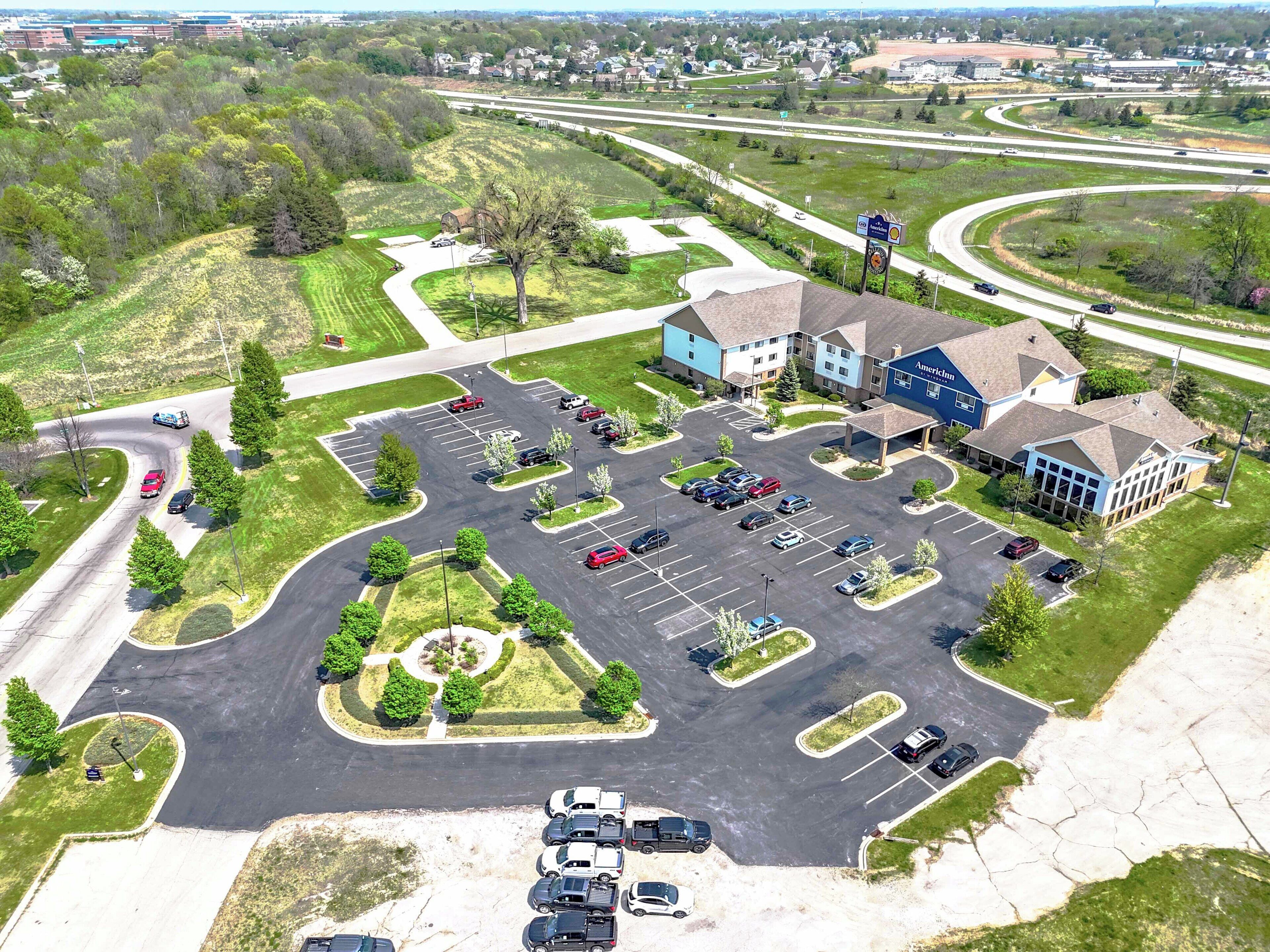 An aerial view of a hotel with a parking lot, a landscaped area, and a highway in the background.