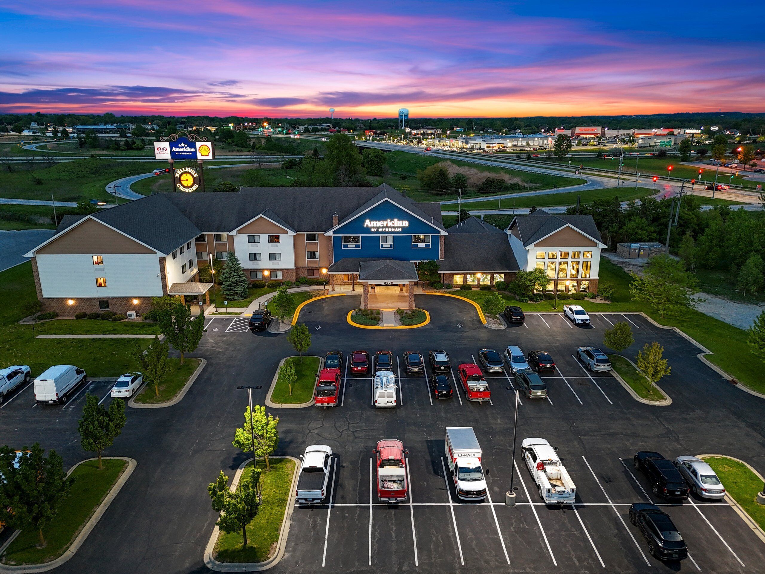 A multi-story hotel with a parking lot filled with various vehicles, surrounded by greenery and a clear sky.