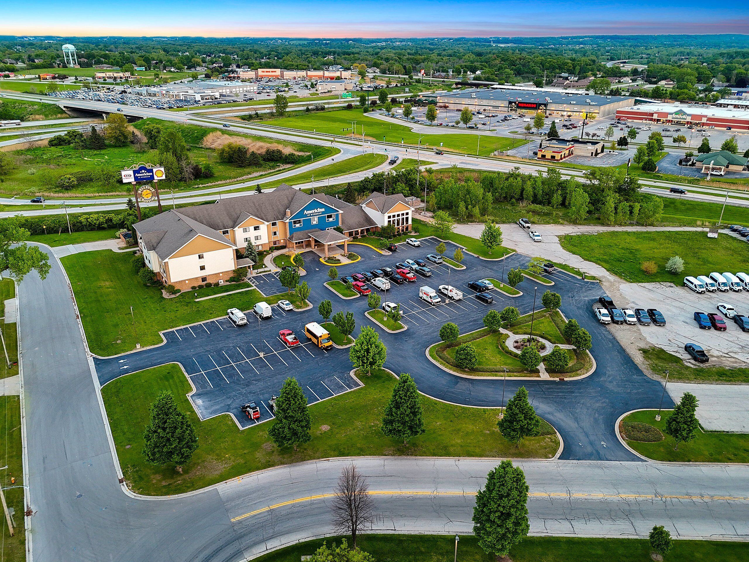 A hotel with a parking lot, a road, and surrounding greenery.