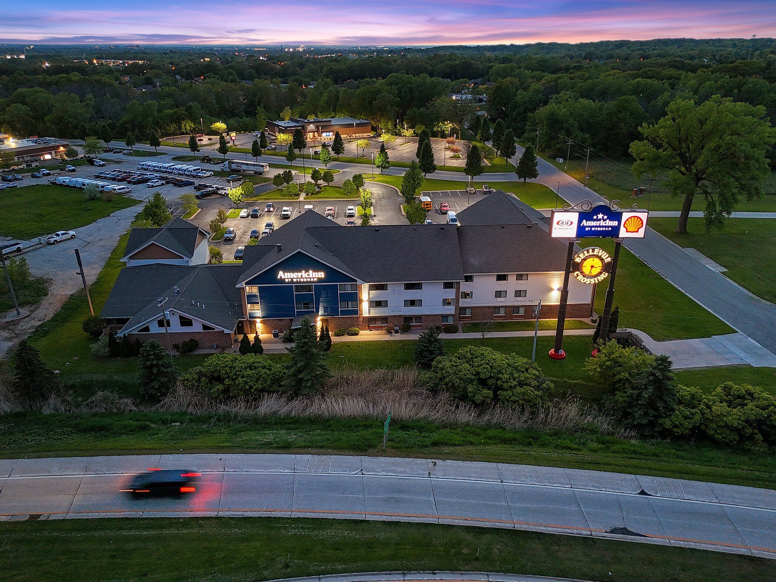 An American Inn by Wyndham with a parking lot, a Shell gas station, and a road with a car passing by.