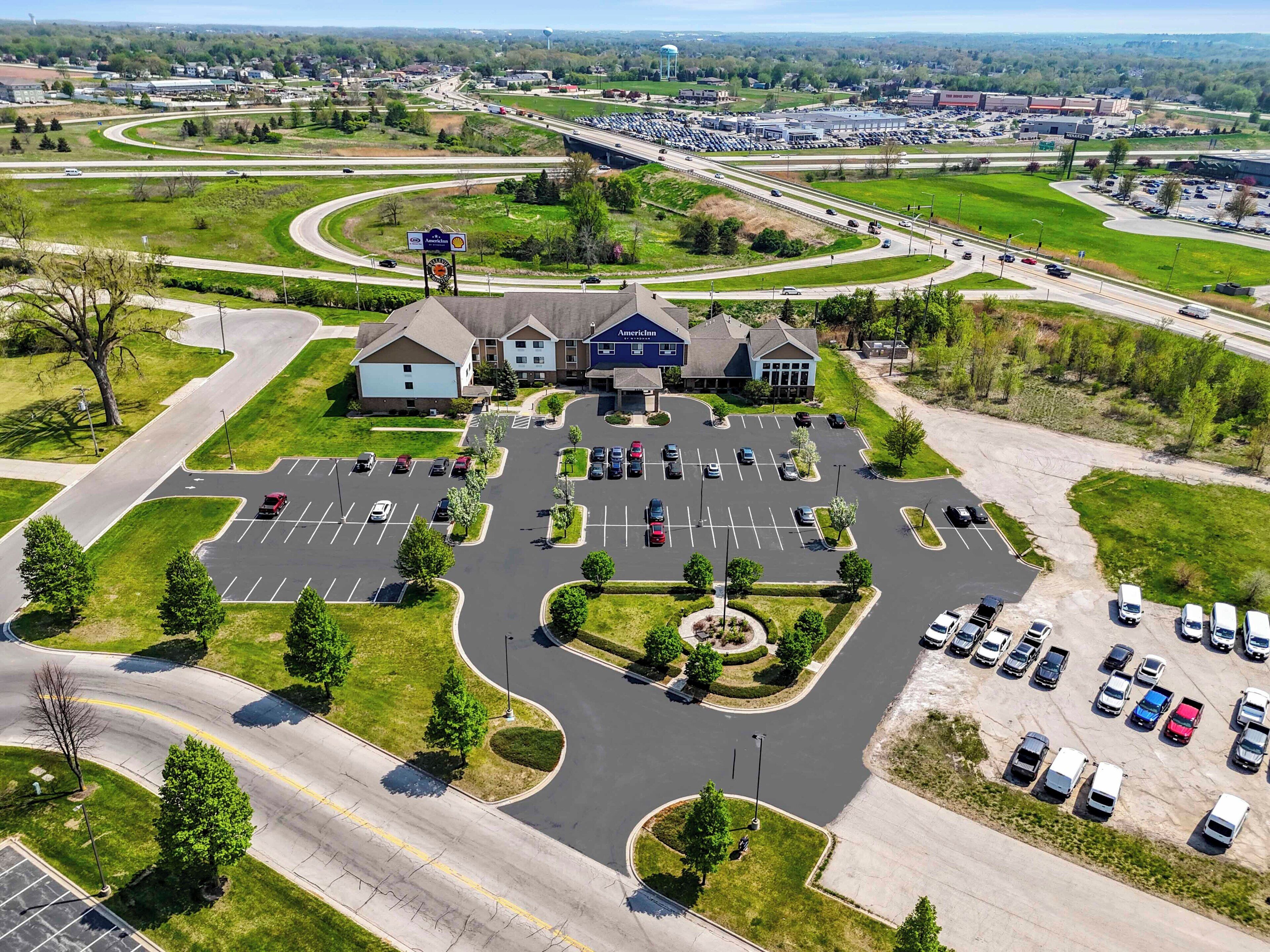 An aerial view of a hotel with a parking lot, surrounded by greenery and a highway.
