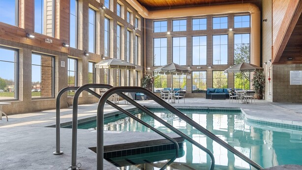 An indoor swimming pool with a metal ladder, large windows, and outdoor seating area. - AmericInn by Wyndham Green Bay East (Green Bay)