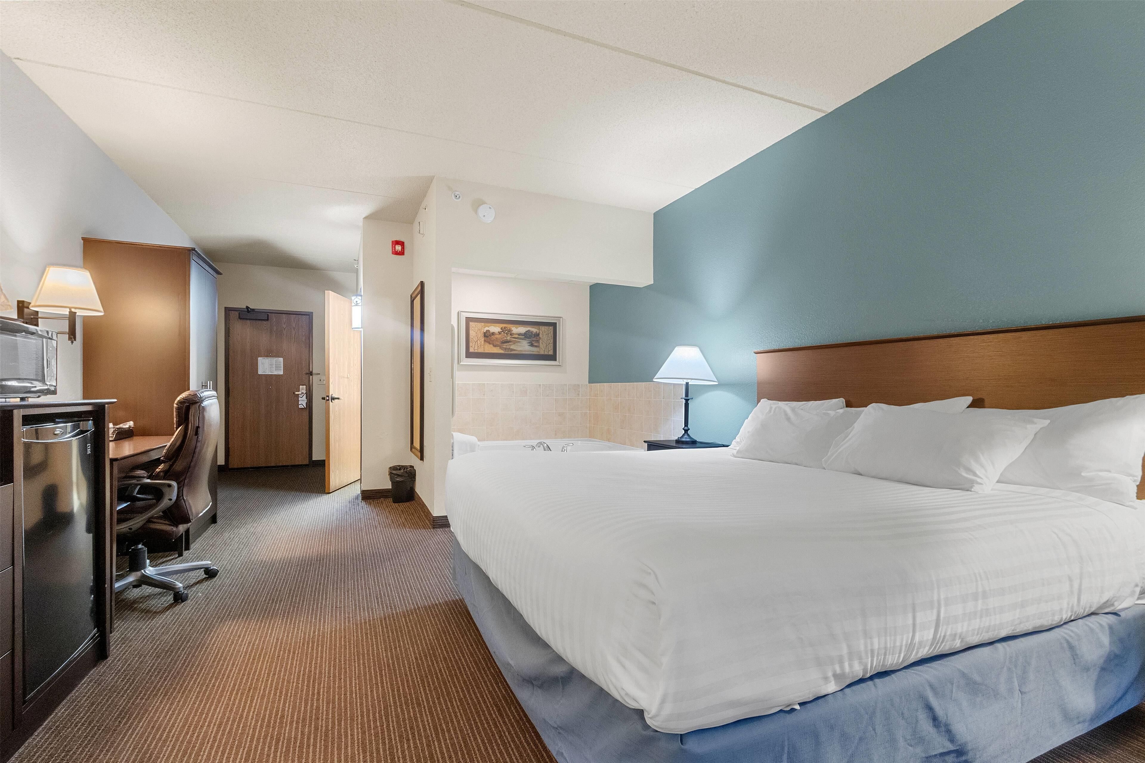 A hotel room with a large bed, a desk with a lamp, a chair, and a small kitchenette.