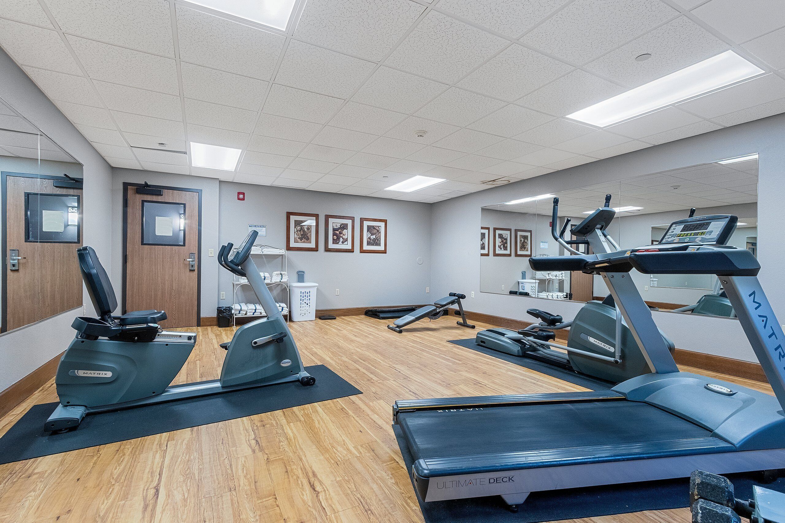 A gym with various exercise equipment, including treadmills and elliptical machines, on a wooden floor.
