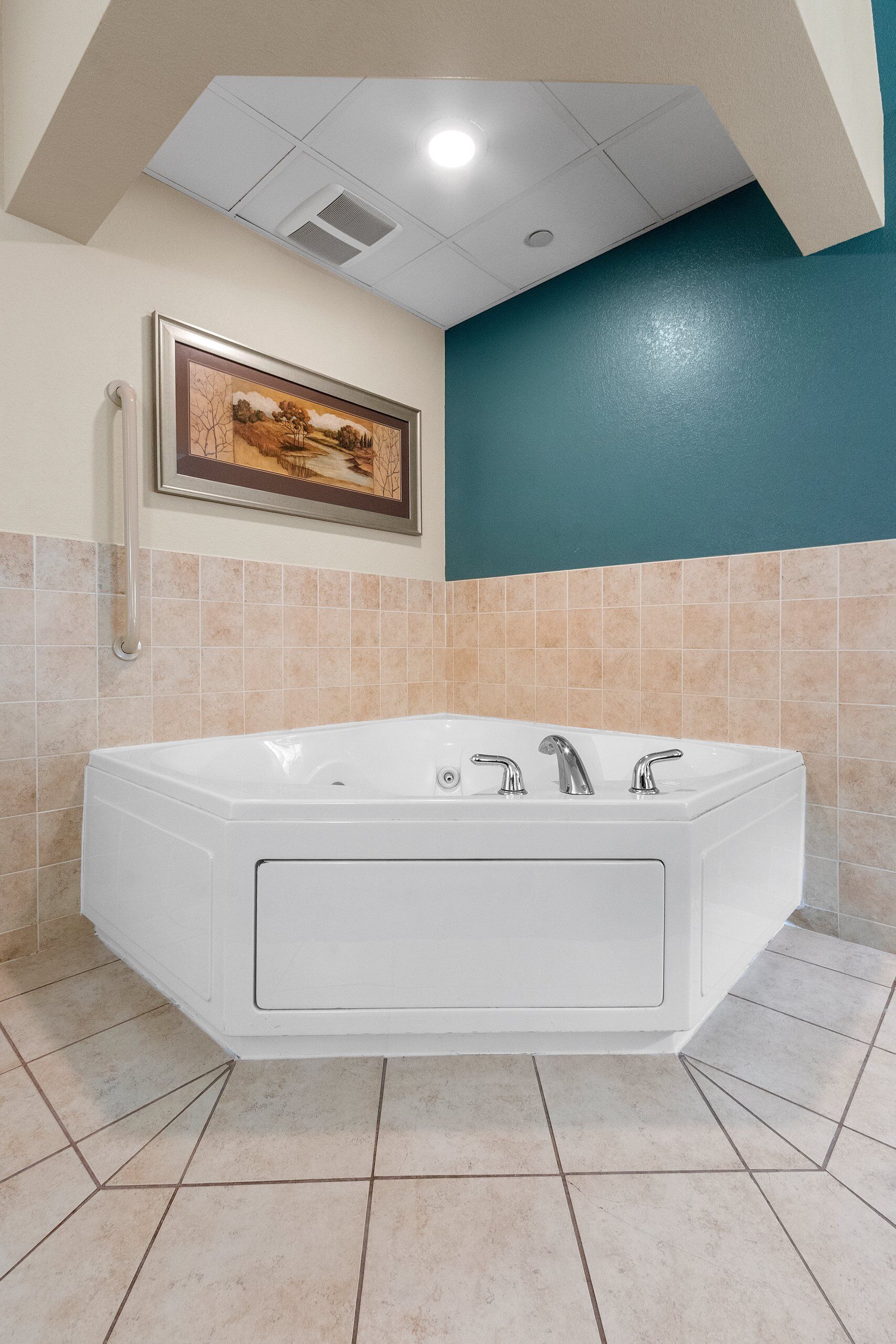 A bathroom with a large bathtub, beige tiles, a teal accent wall, and a framed picture above the tub.