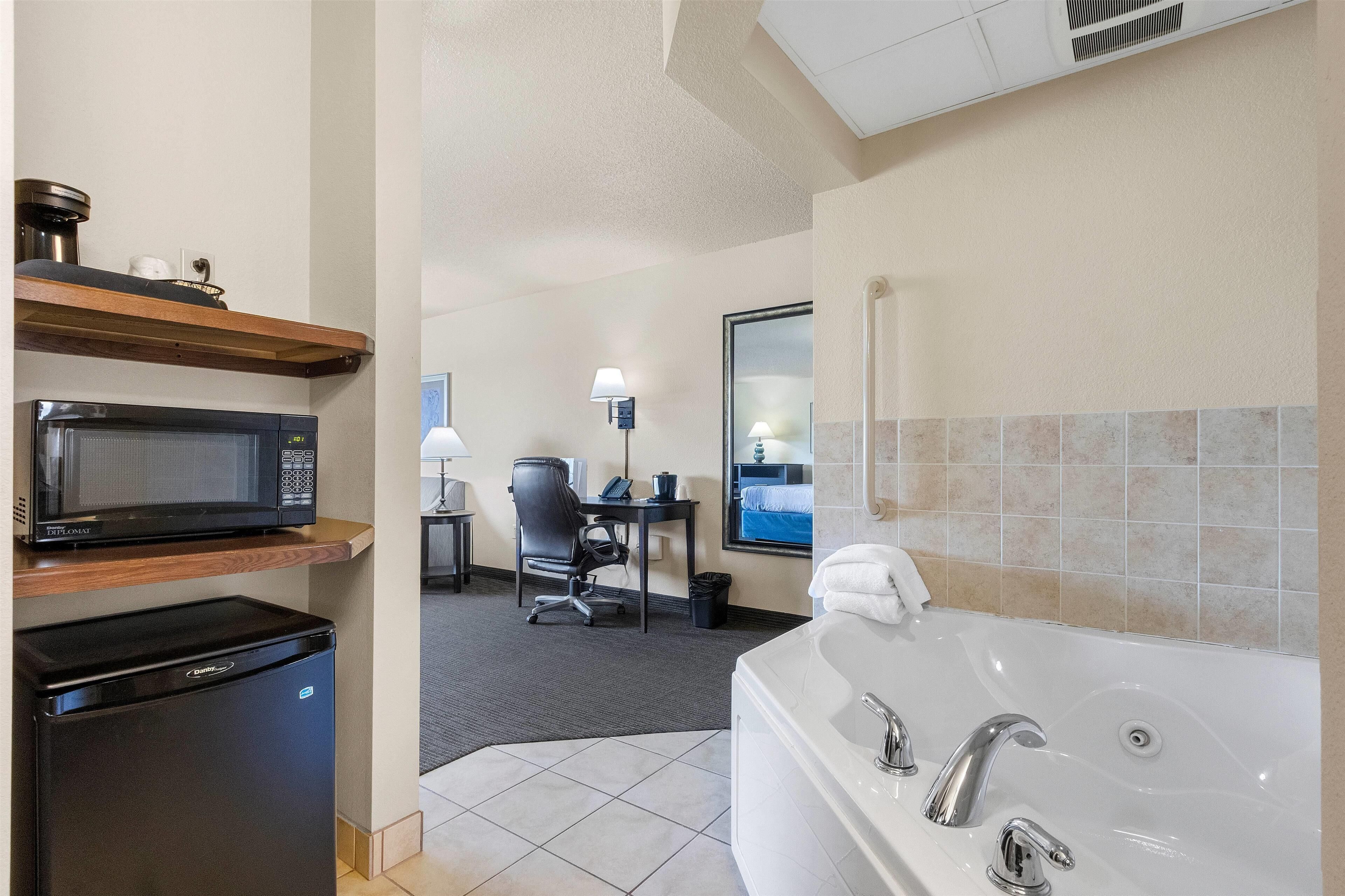 Un baño de hotel con una bañera blanca, microondas, refrigerador, escritorio con silla y espejo.