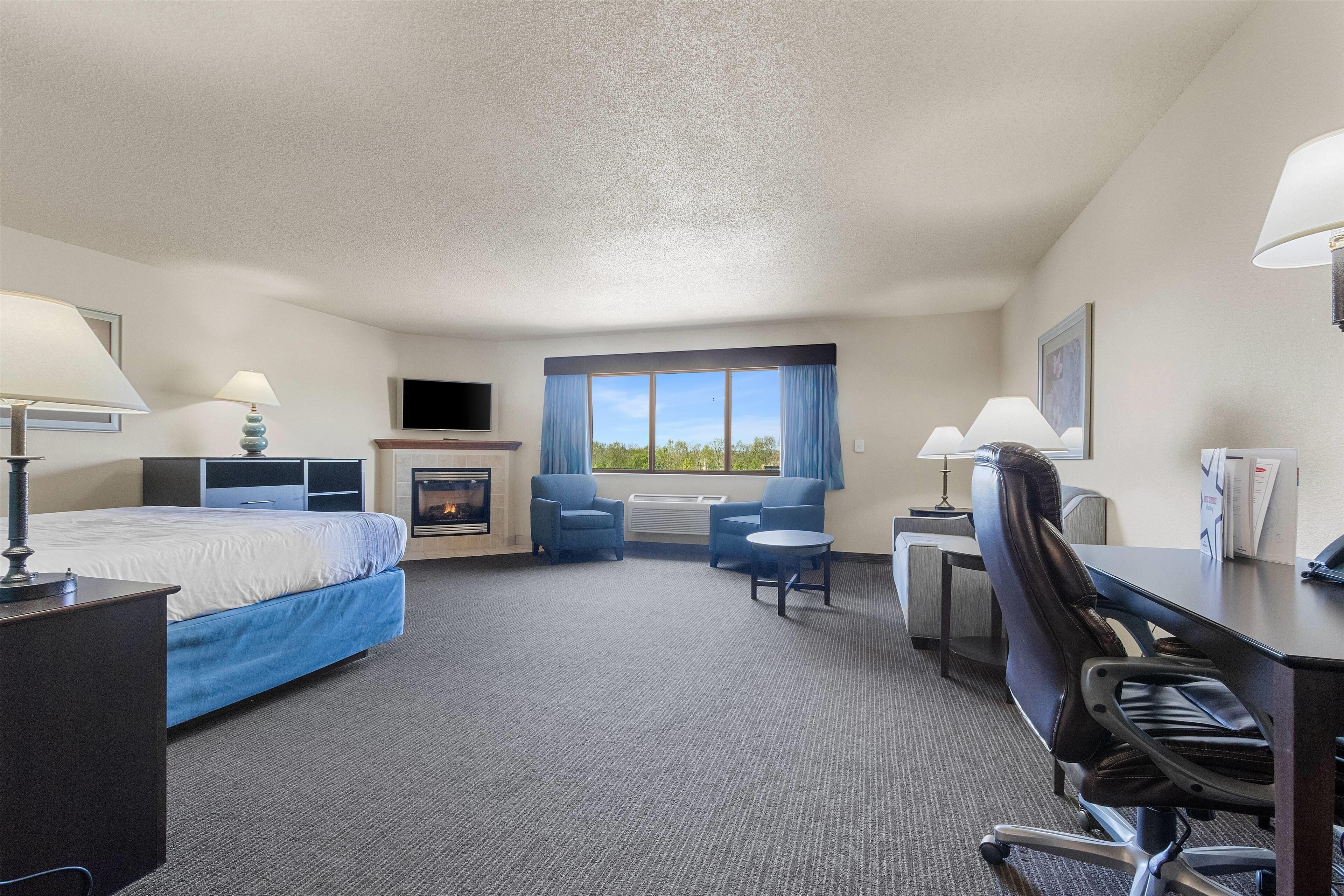 A hotel room with a bed, desk, chairs, a fireplace, and a large window with curtains.