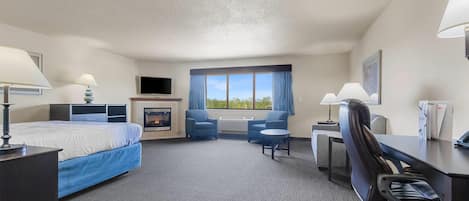 Una habitación de hotel con cama, escritorio, sillas, chimenea y un ventanal con cortinas.