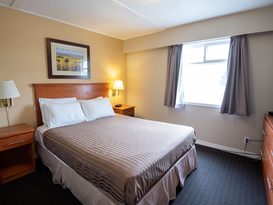Ein Hotelzimmer mit einem großen Bett, einem Holznachttisch, einer Kommode, einem Fernseher, einem Fenster mit Vorhängen und einem an der Wand hängenden Bild.