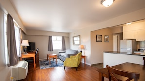 A modern living room with a wooden floor, a sofa, a yellow armchair, a dining table, and a kitchen area with a refrigerator. - Canadas Best Value Inn Prince George (Prince George)