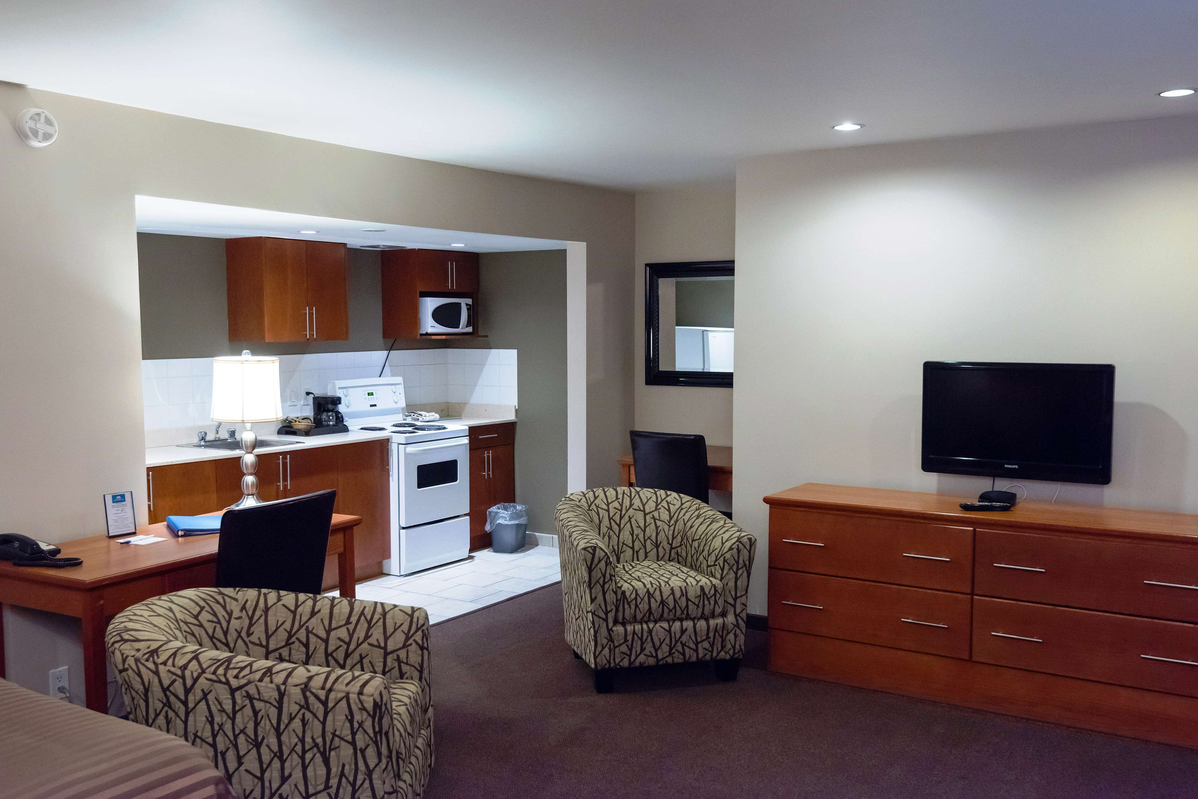 Una habitación de hotel compacta con kitchenette, televisor, escritorio con silla y sillón estampado.