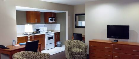 Una habitación de hotel compacta con kitchenette, televisor, escritorio con silla y sillón estampado.