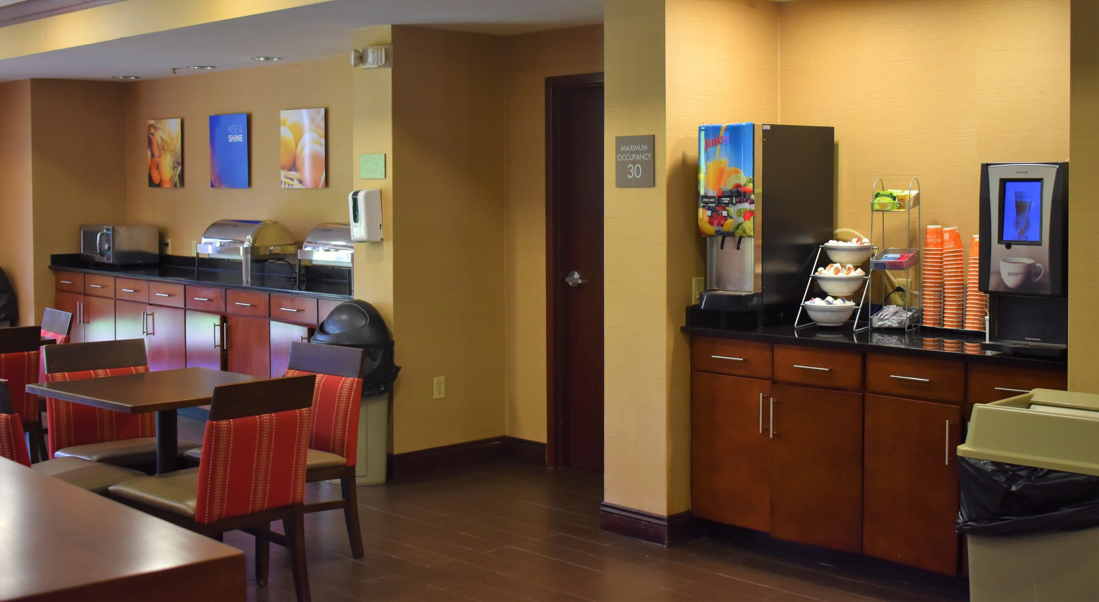 A hotel room with a kitchenette, dining area, and a refrigerator with a juice dispenser.