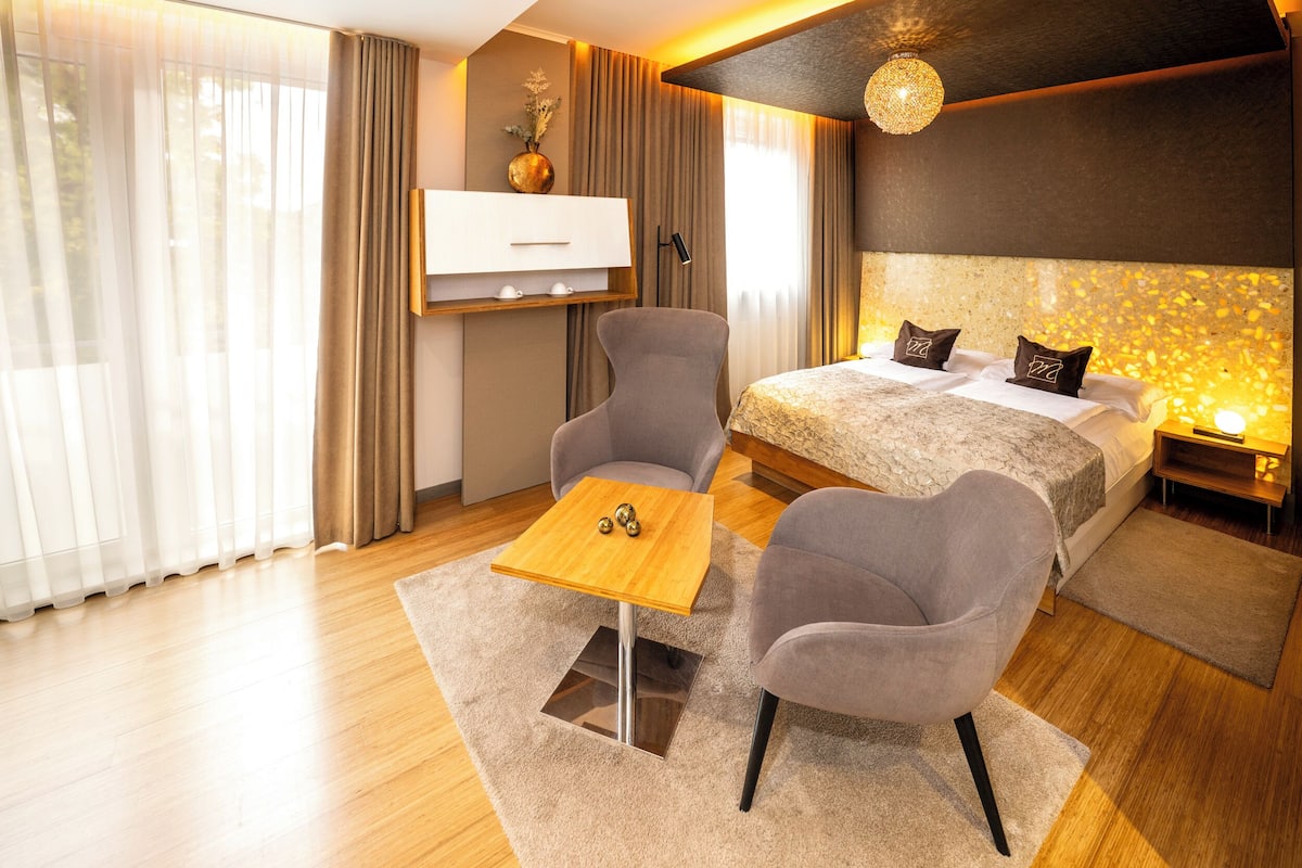 Una habitación de hotel moderna con una cama, dos sillones, una mesita de madera, un televisor y un detalle decorativo en la pared.