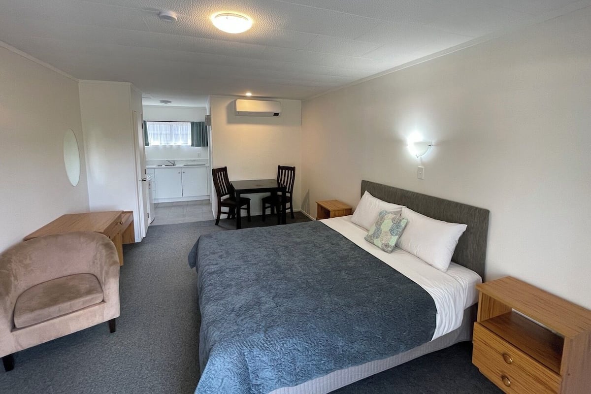 A compact hotel room with a bed, a small desk, a chair, and a kitchenette in the background.