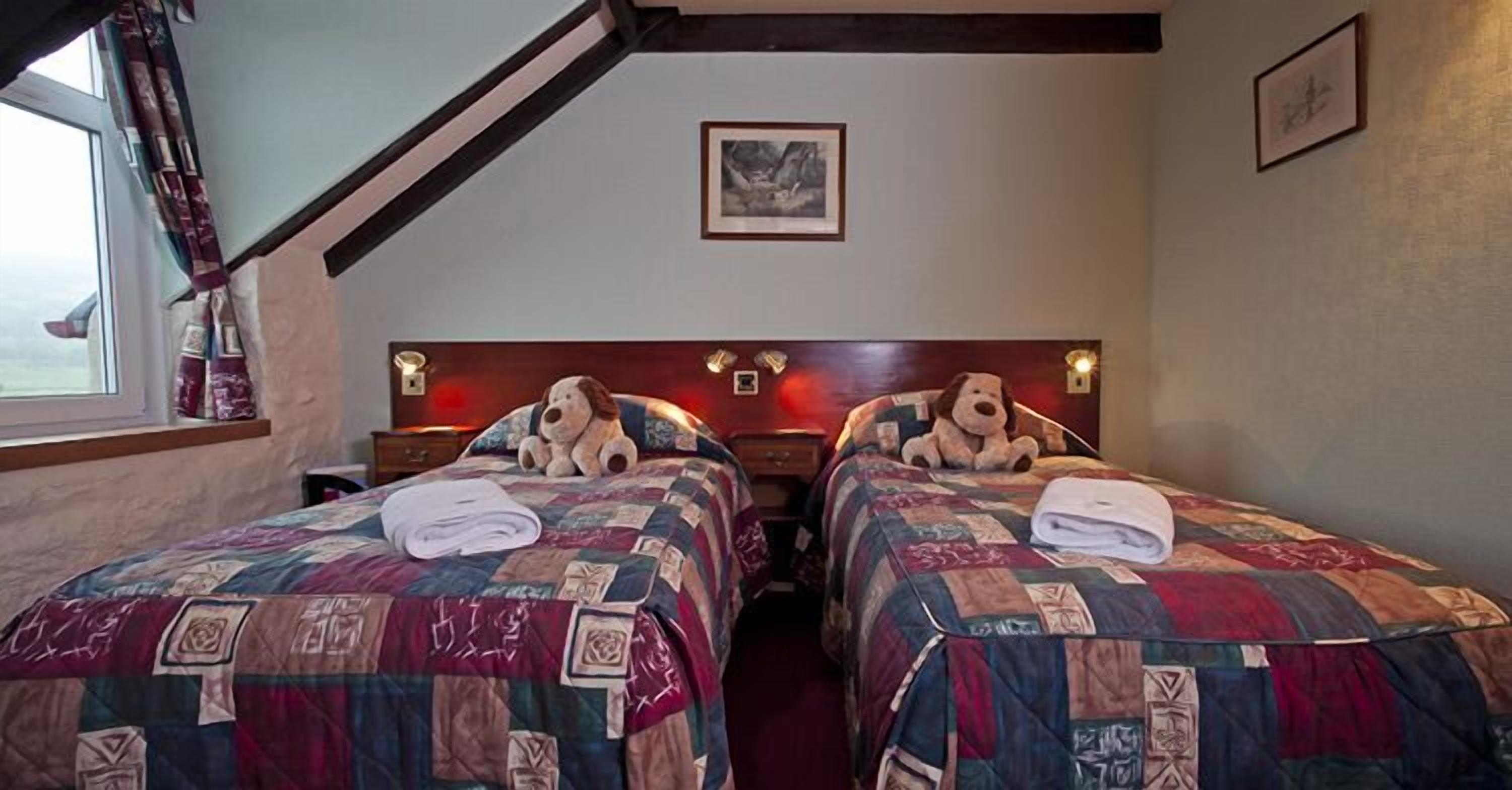 A room with two beds, each with a quilted bedspread and a stuffed toy on top. There is a window with curtains, a framed picture on the wall, and a headboard with lights.