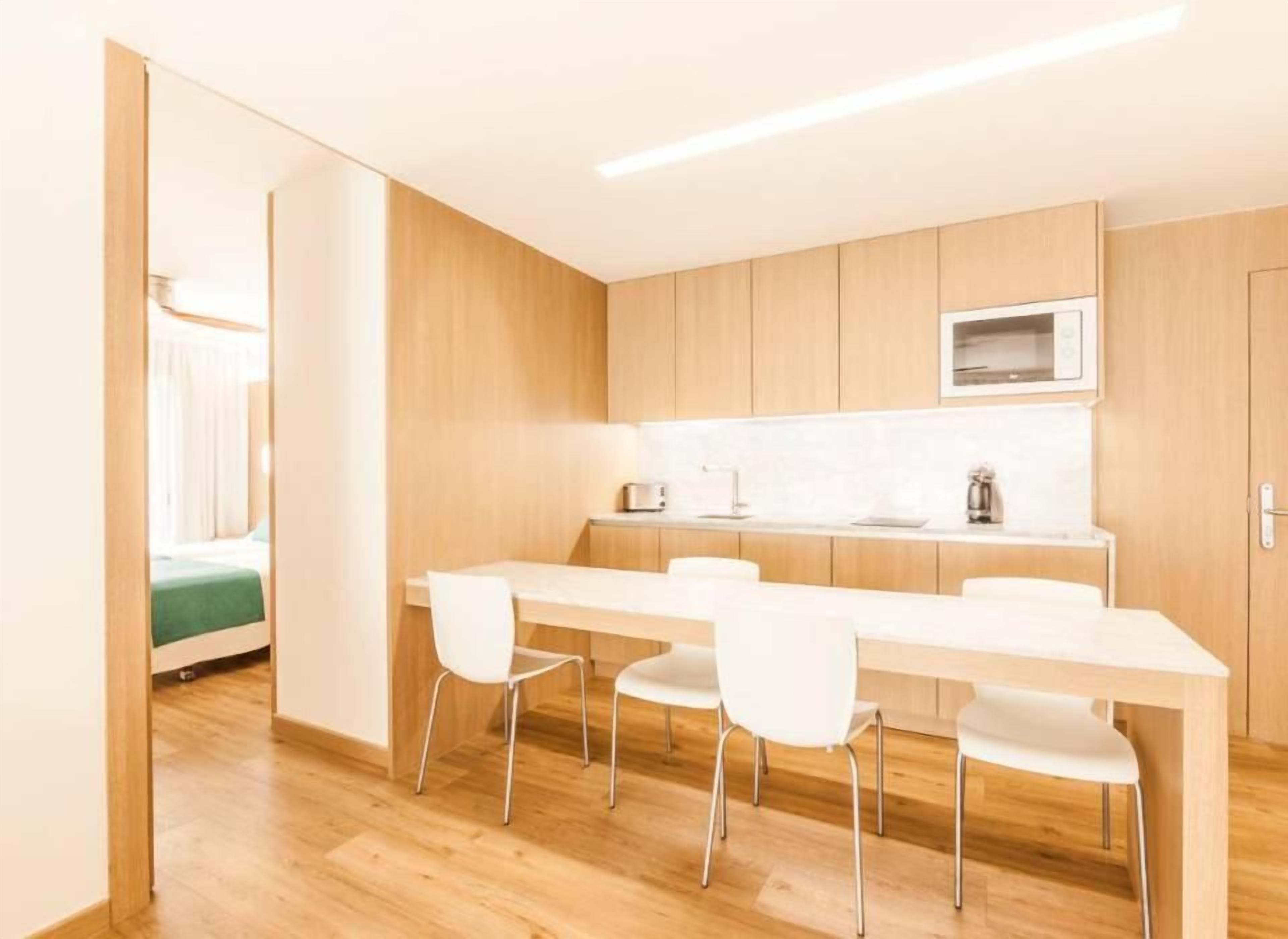 A modern kitchen with a white countertop, wooden cabinets, and a microwave. There are four white chairs around the table.