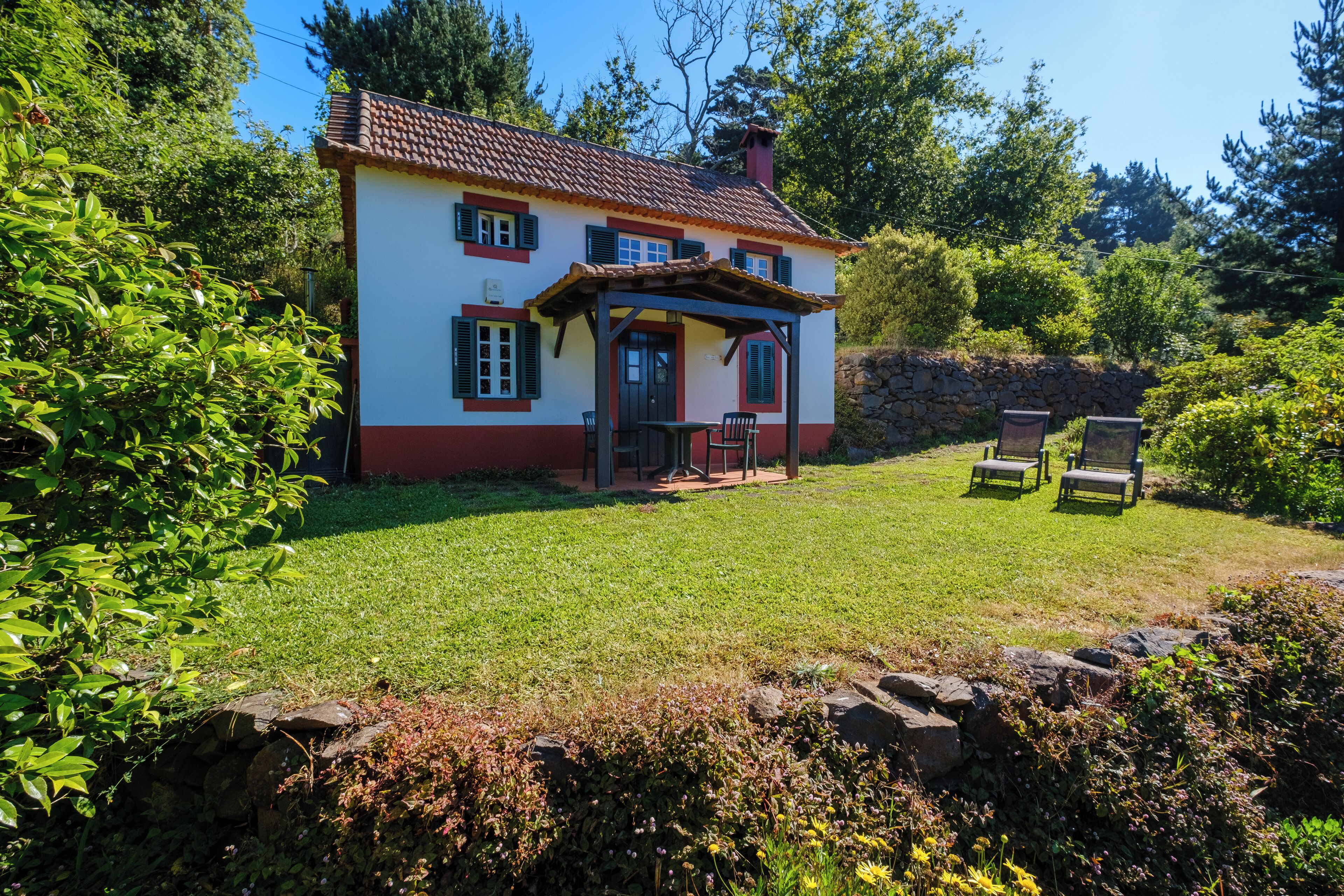 A house with a tiled roof, a porch with chairs, and a stone wall in the background.