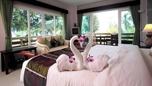 Ein Hotelzimmer mit einem großen Bett, zwei schwanenförmigen Kissen, einem Fernseher und einem Balkon mit Blick auf Grünflächen.