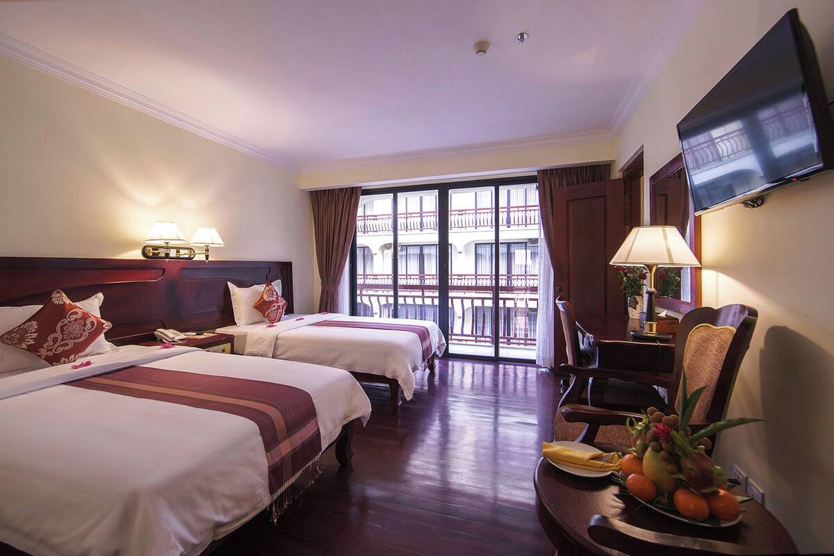 A hotel room with two beds, a TV, a desk with a lamp, and a balcony with a view.