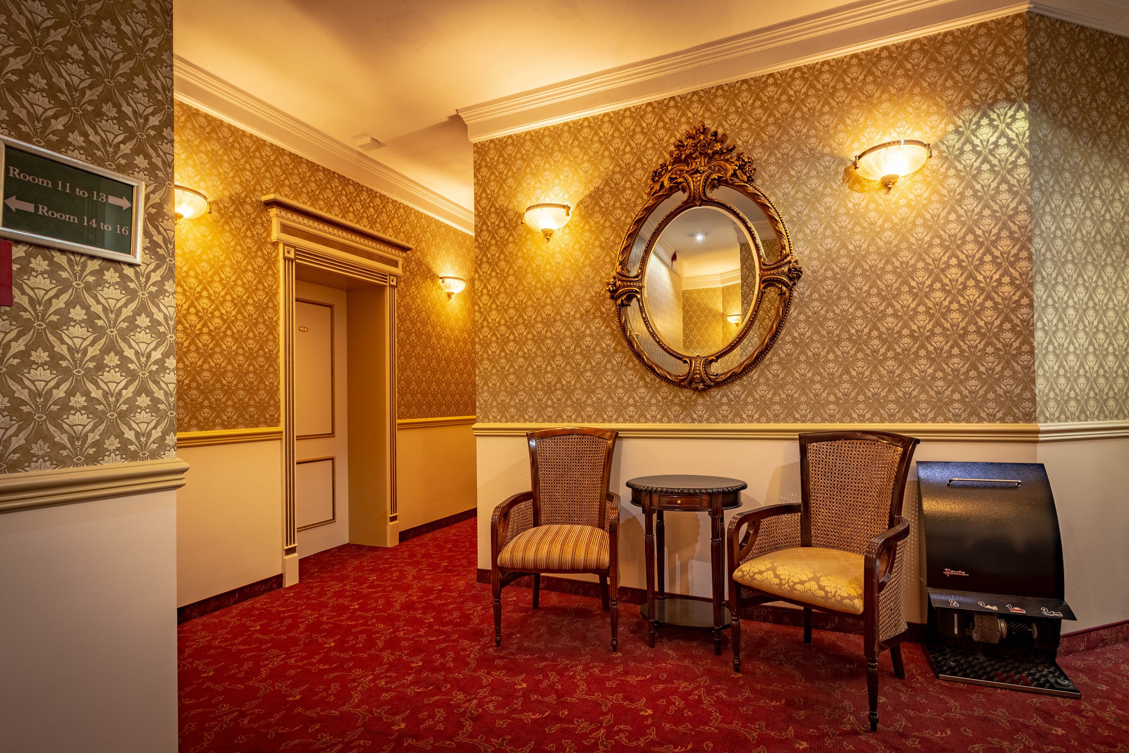 A hotel hallway with patterned wallpaper, a mirror, two chairs, a small table, and a grill.