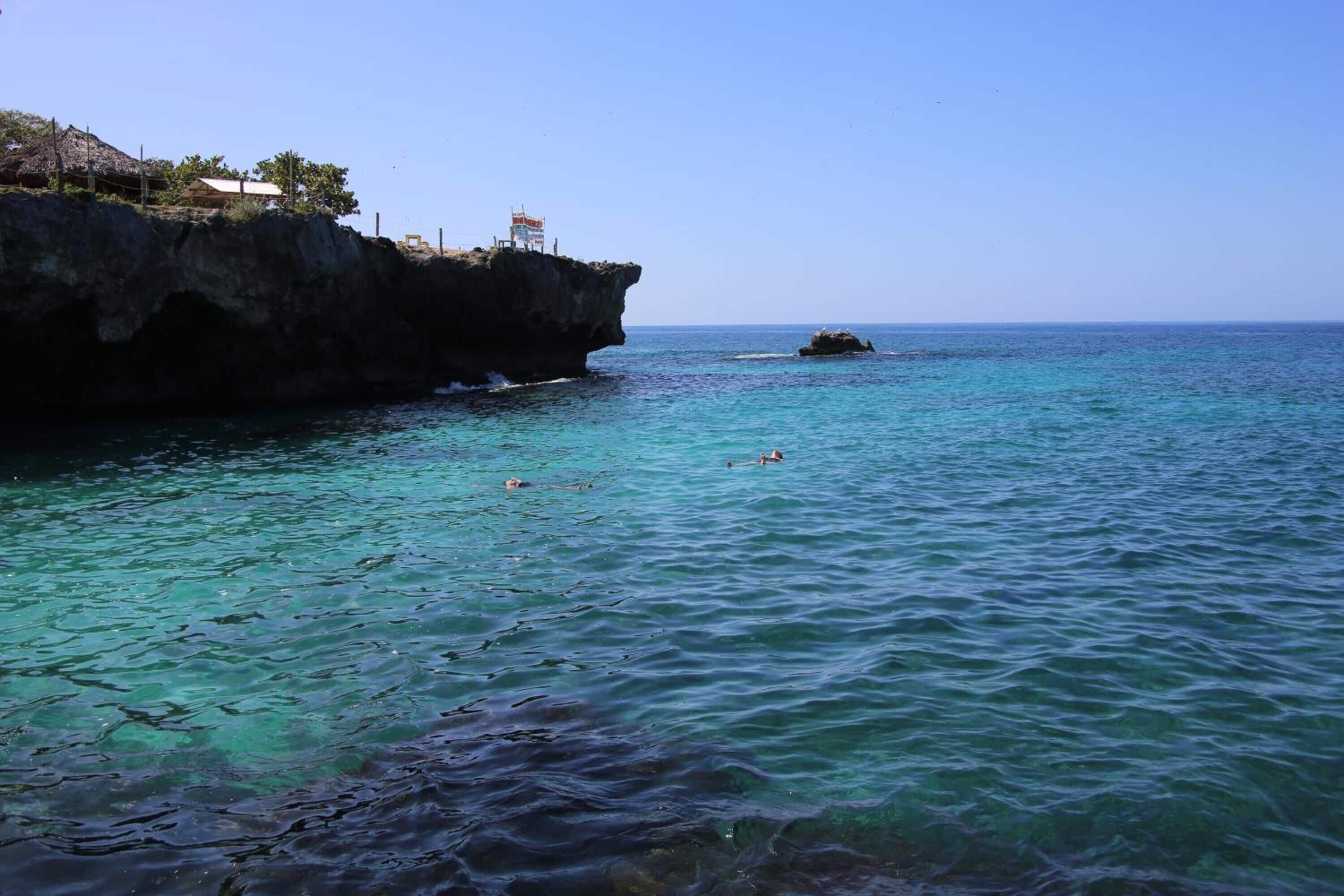 A coastal scene with clear turquoise water, a rocky cliff, and a few people swimming.