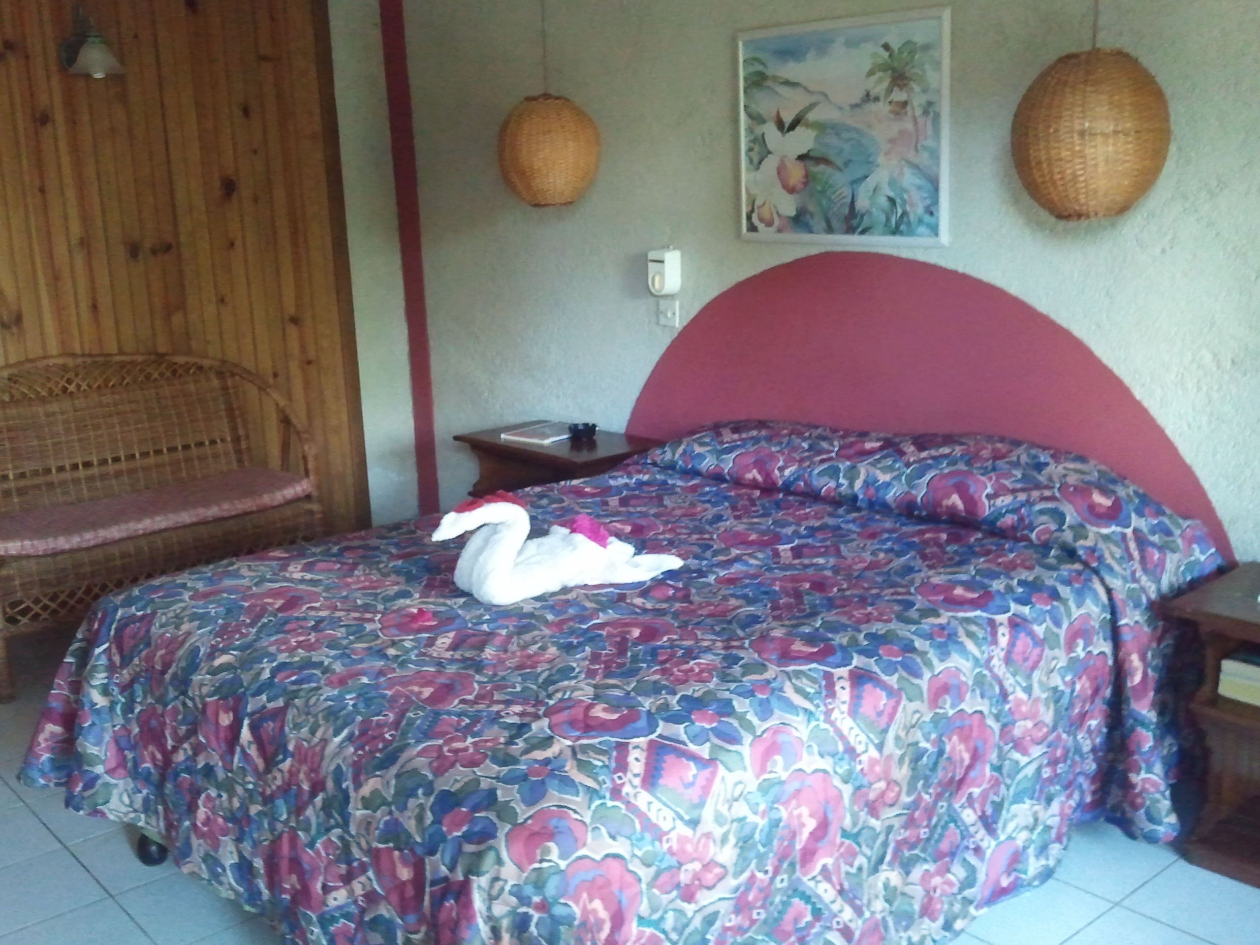 Una stanza con un letto, una sedia in vimini, un comodino e un quadro appeso al muro.